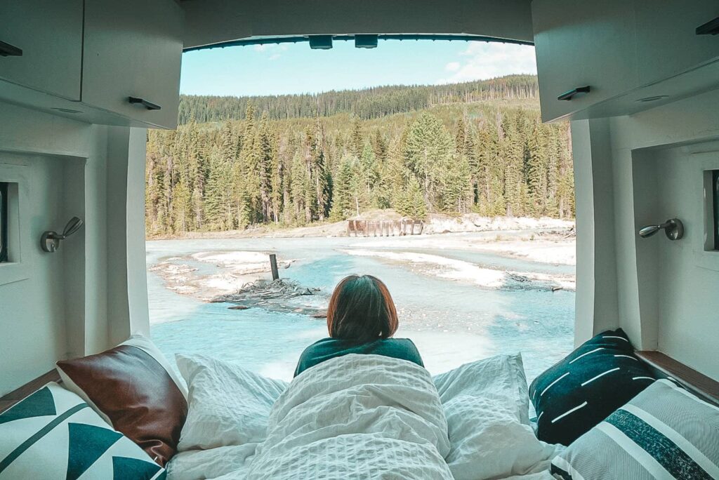 Woman laying on the bed inside a camper van looking out the back doors towards a river and trees