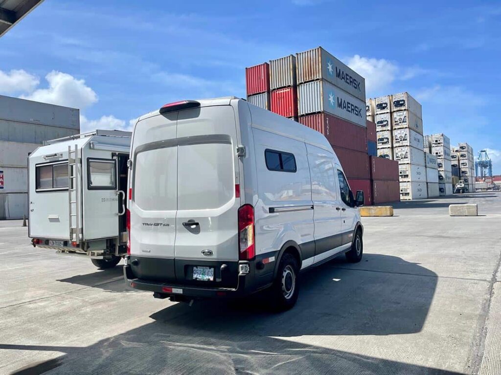 Two camper vans parked at a shipping port with shipping containers in the background