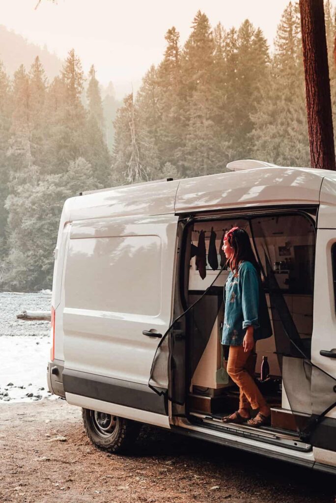 Yuko standing inside a camper van and looking out across the river and forest
